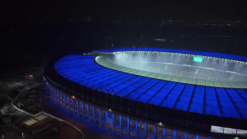 An aerial view of a large sports stadium at night illuminated by blue exterior lights
