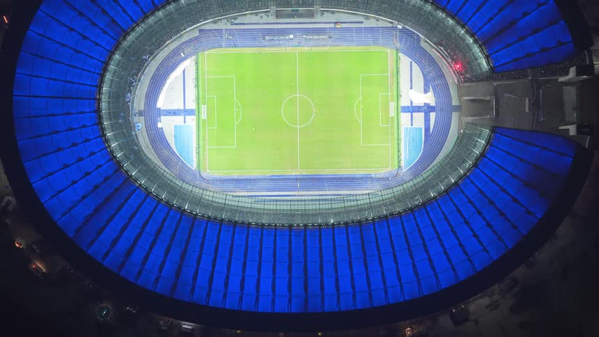 An aerial drone shot captures a professional soccer stadium at night with its blue roof illuminated
