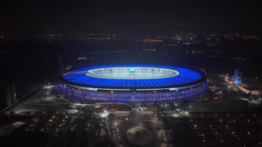 A stadium with its roof glowing in vibrant blue lights while the surrounding urban landscape of parking lots and roads sits in darkness under a clear sky
