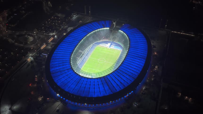 Aerial view of a brightly illuminated football stadium with a glowing blue roof at night