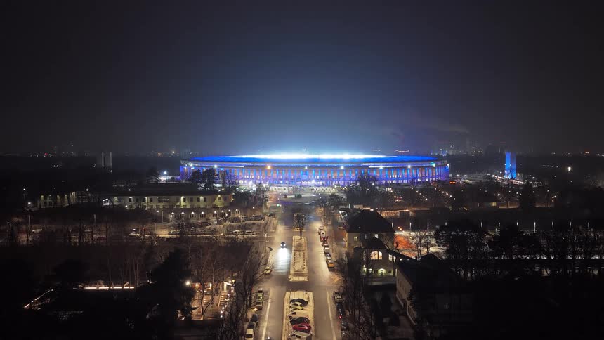 A brightly lit stadium glows blue at night while a line of car headlights forms visible traffic along the central road leading toward it