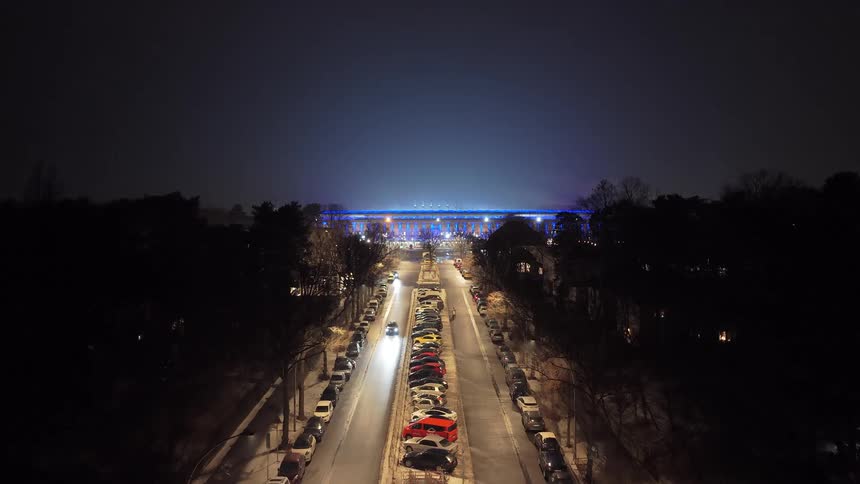 A high view looking down a long city street at night with parked cars and moving headlights leading toward a large sports stadium with a glowing blue roof and bright lights under a dark sky
