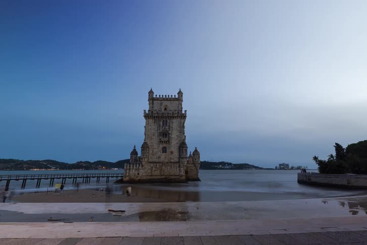 People sit on the waterfront in the foreground looking out toward a historic stone tower standing alone in the calm water under a pale evening sky