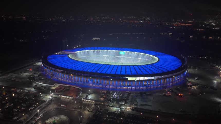 A football stadium lit up at night with the roof illuminated in blue light in the middle of the city