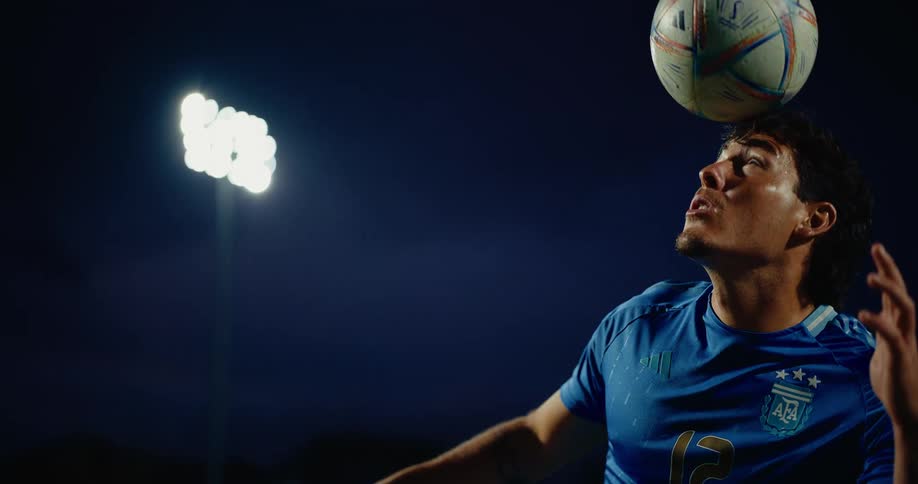 A soccer player wearing a blue jersey hits a ball with his head during a night session at a stadium while bright floodlights shine in the dark background
