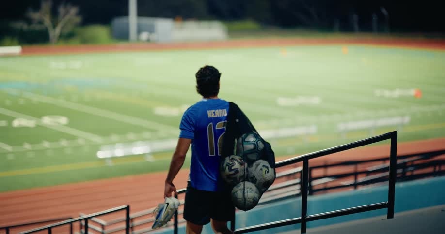 A soccer player walks down the bleachers toward an empty field at night carrying a mesh bag full of ballshe holds a pair of cleats in his hand as he moves away from the camera toward the illuminated turf
