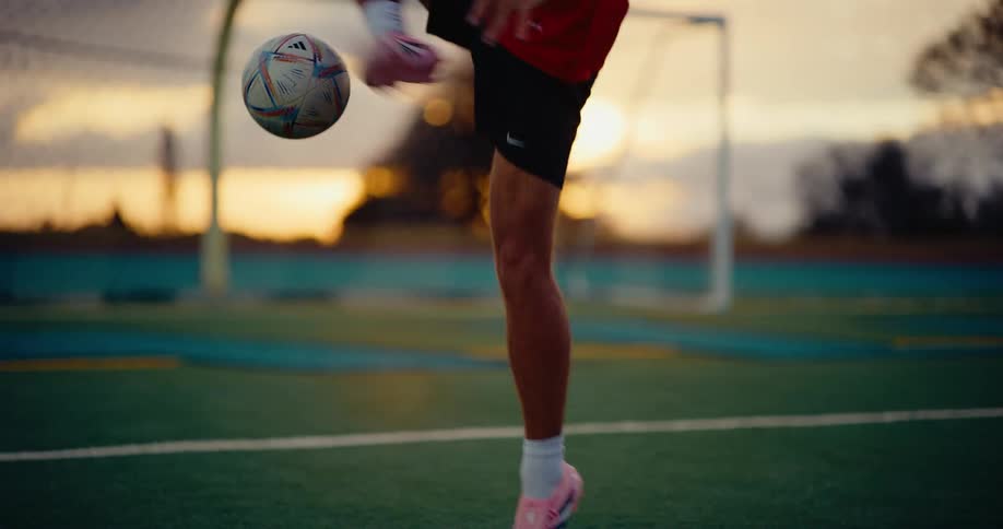 A man wearing a red sports jersey uses his feet and knees to keep a soccer ball in the air on a green field during a sunset with a goal post in the background