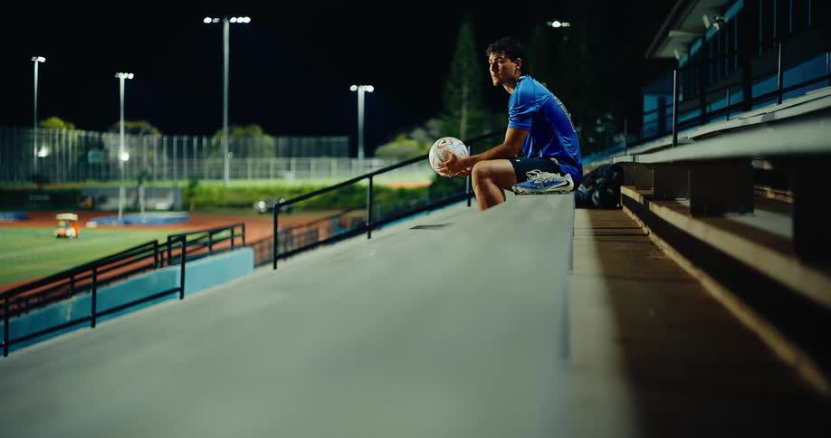 A man in a blue sports shirt sits alone on stadium bleachers at night holding a soccer ball while bright field lights shine in the background