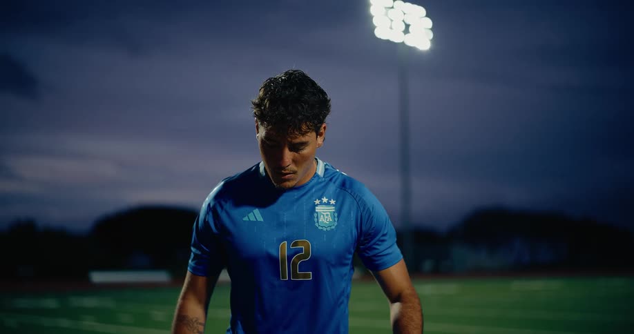 A man wearing a blue sports jersey uses his feet and knees to keep a soccer ball in the air on a green field at night under bright stadium lights