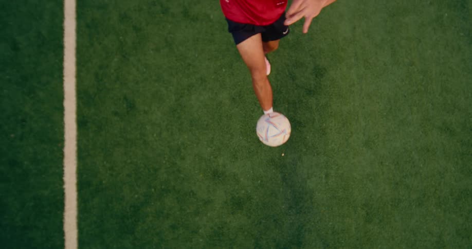 A man wearing a red shirt and black shorts juggles a soccer ball with his feet on a green turf field marked by a white sideline
