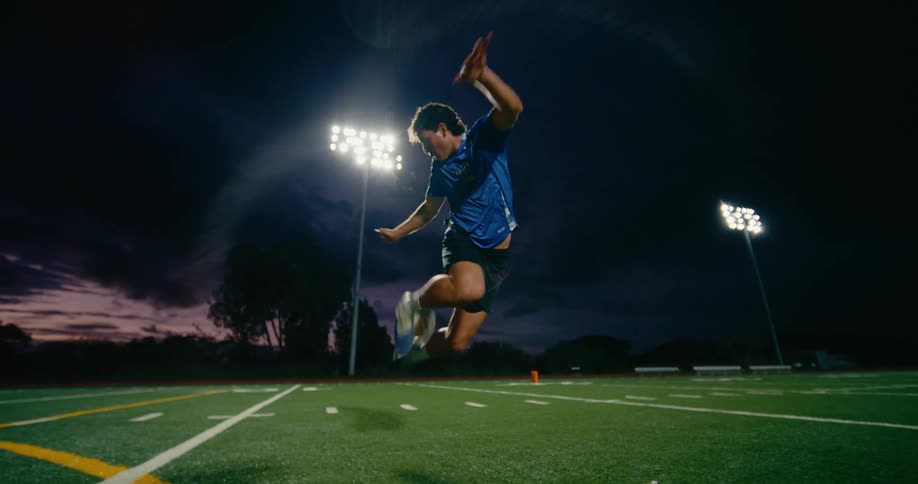A male soccer player wearing a blue jersey juggles a ball with his feet and knees on a green turf field under bright stadium lights at night while the sun sets in the background