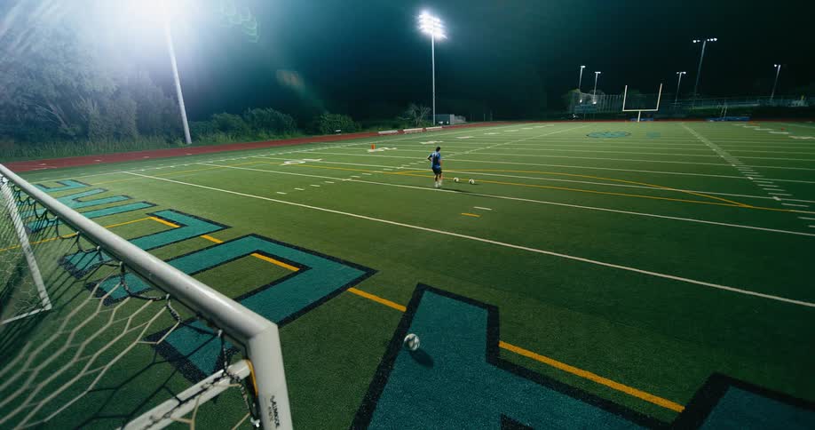 A soccer player practices kicking a ball on a lit field at night