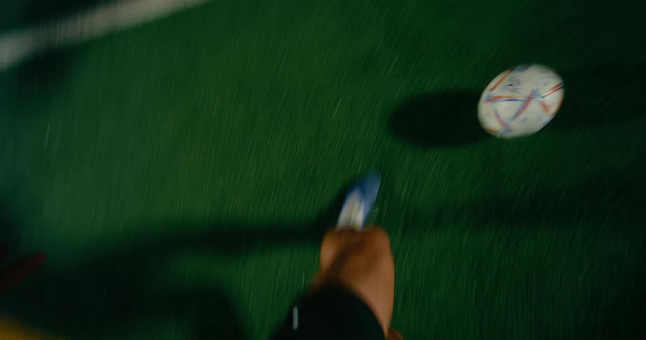 A view from the perspective of a player shows their feet and a soccer ball as they dribble across a green field at night while another player in a red shirt tries to defend