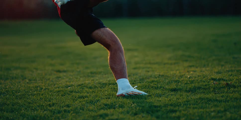 A male soccer player kicks a ball across a green grass field during sunset capturing his legs and white cleats in a dynamic athletic motion while the background remains softly blurred
