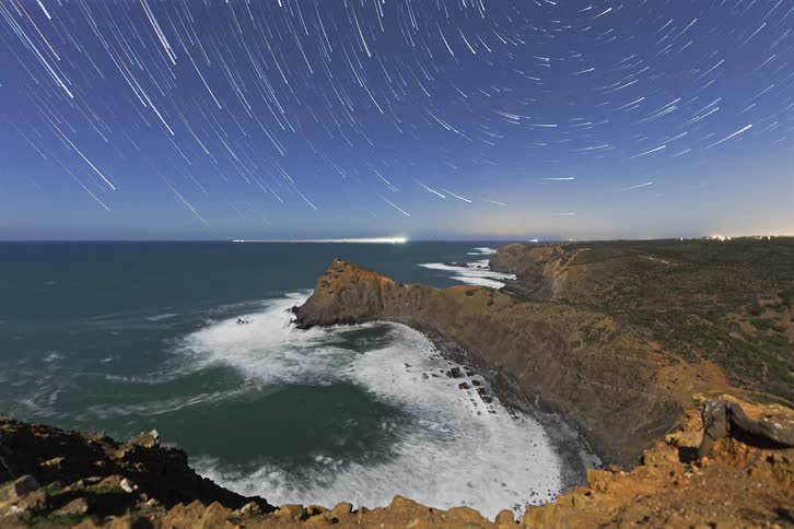 Mountains and cliffs along the coast overlooking the water as waves roll into shore and boats sail with stars glowing in the sky