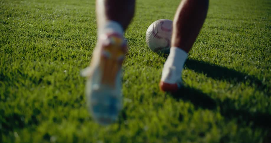 A young soccer player wearing a red jersey with the name ronaldo on the back practices dribbling on a sunny grass field the camera captures closeup shots of his feet and the ball as he moves across the pitch toward a tall fence