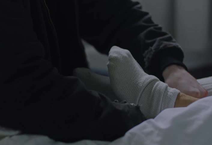 A young man sits at a bedside helping an elderly person put on socks