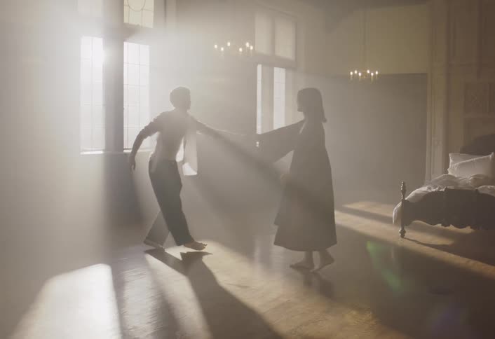 A man and woman dance together in a sunlit bedroom filled with hazy light from large windows