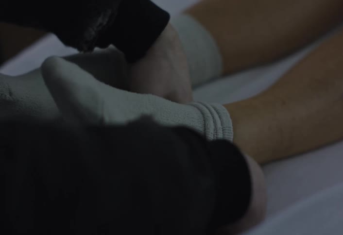 A person lying on a bed wearing socks while another person removes one of the socks