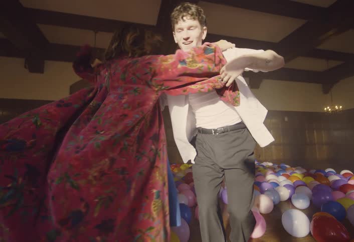 Balloons cover the floor as a young man and woman dance together with sunlight shining brightly in the windows