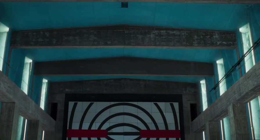 A view inside a large concrete building with a blue ceiling shows a wall with a target pattern of black and white circles and a red line in the middle