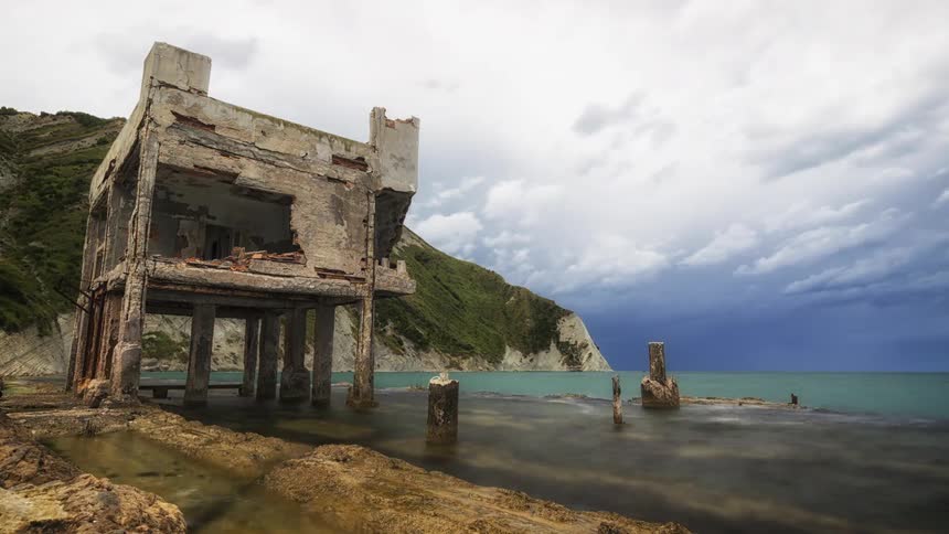 An abandoned building on stilts stands in shallow coastal water beside rocky cliffs under cloudy skies