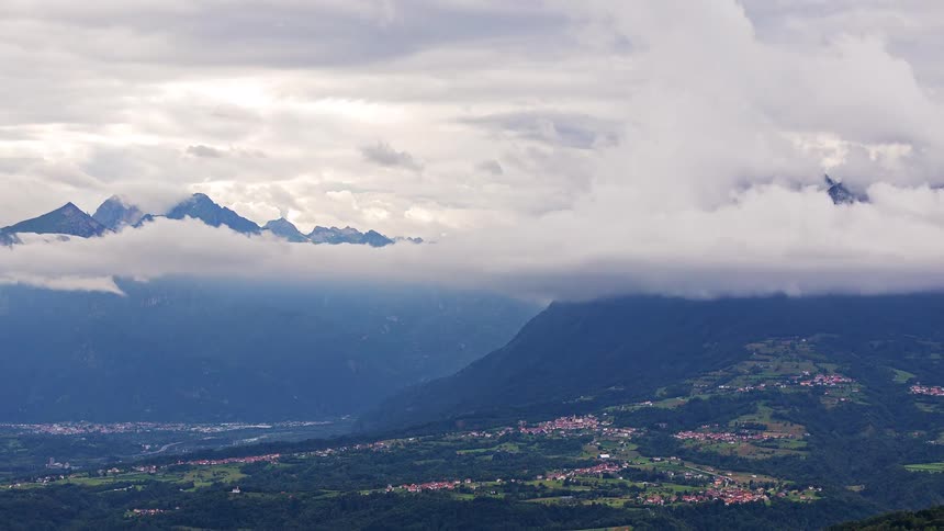 Low clouds drift across a mountain range overlooking a green valley with small villages under an overcast sky