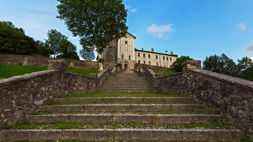 A view from the bottom of a stone staircase shows a large old church and building on a hill with green grass and trees under a blue sky