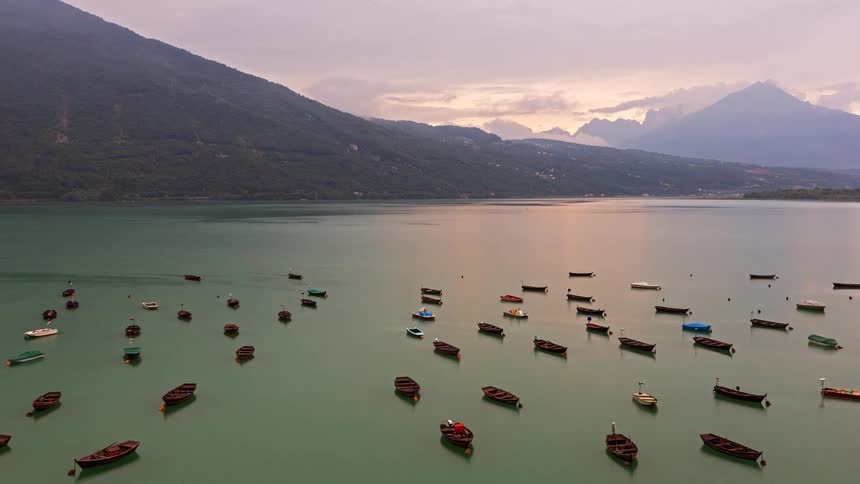 Dozens of small boats float on a calm lake surrounded by misty mountains under a cloudy sky