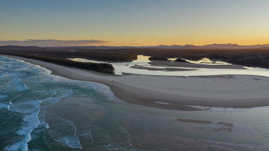 An aerial view shows a wide sandy beach and a river that flows into the ocean while the sun sets over the distant hills