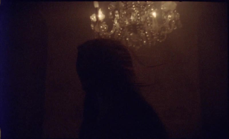 A young woman in a dim windy chandelierlit room looks ahead with a tense mysterious expression as warm shadows surround her