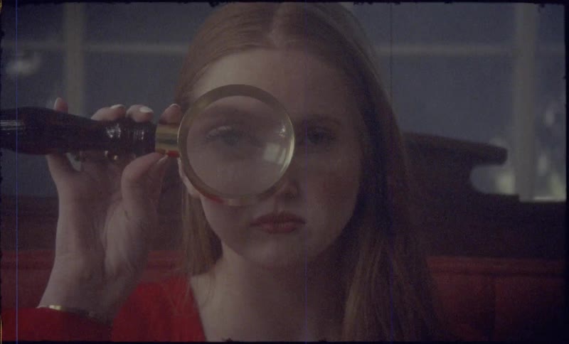 A woman with red hair looks through a magnifying glass directly into the camera lens
