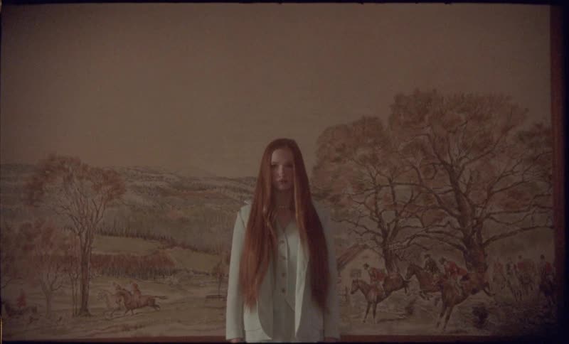 A view shows a woman with long red hair as she stands still in a room in front of a large painting on a wood wall