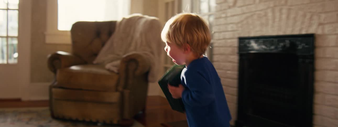 A small boy in a blue sweater runs through a sunlit living room while clutching a green bag of coffee