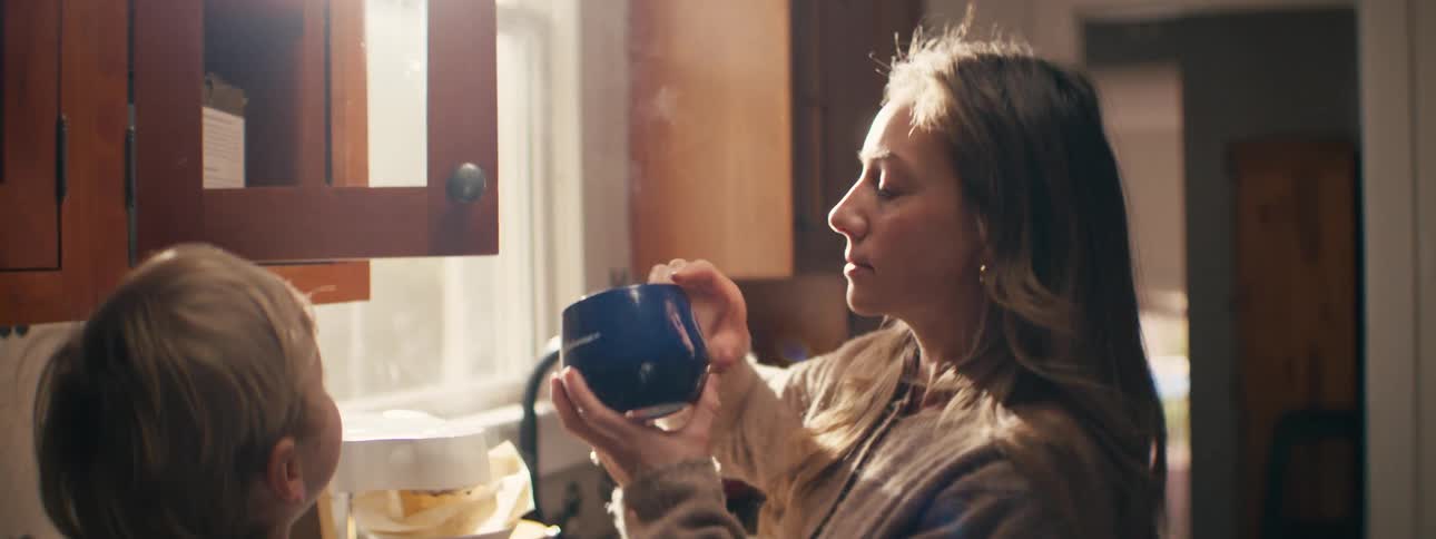 A woman stands in a sunlit kitchen grabbing a mug out of the cupboard to hand to a small boy next to her