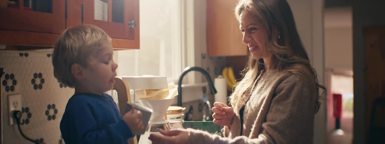 A mother hands her son a cup as he sneezes in the kitchen