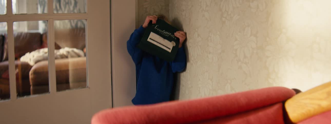 A young boy in a blue sweater runs through a living room and hides behind a chair while holding a bag of coffee over his face