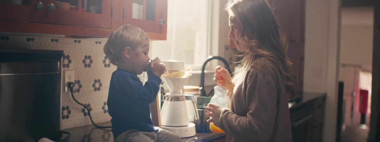 A woman in a warmly lit kitchen helps a young child hold a small cup while preparing a drink to give to him beside the counter