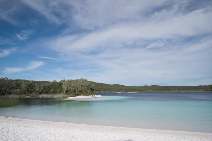 This timelapse captures wispy white clouds moving across a bright blue sky over a serene turquoise lake the scene features a pristine white sand beach in the foreground and a lush forested shoreline in the distance