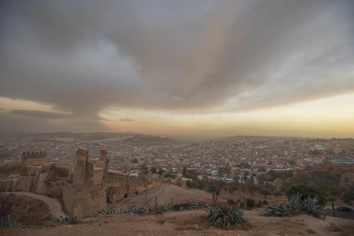 People walk on a hill near a ruin at the edge of a city at nightfall