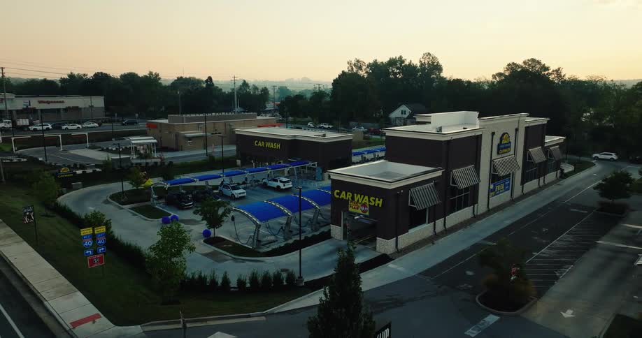 A modern car wash facility and adjacent commercial buildings at dusk showing a parking lot with blue vacuum stations surrounding greenery and a distant pharmacy under a hazy evening sky