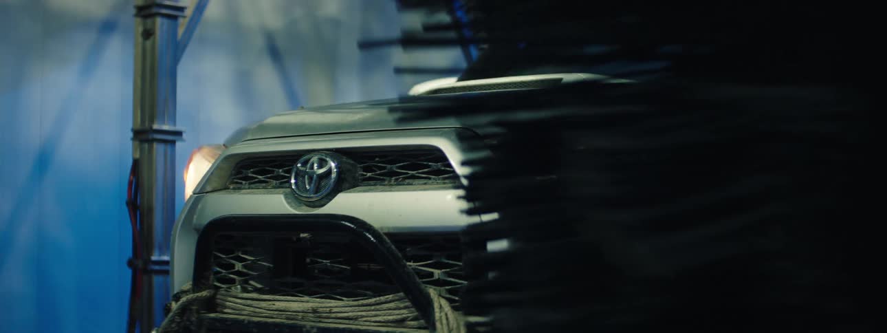 A white toyota vehicle with its left headlight on is partially obscured as a large dark rotating carwash brush sweeps across the frame inside an automated wash bay