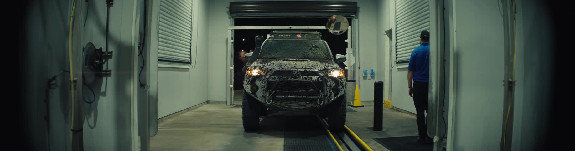 Awhite toyota suv with its headlights on moves forward along the track inside a brightly lit automated car wash bay with foam and grime covering the front grille and bumper being directed by a worker