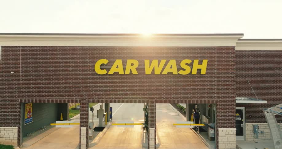 An aerial shot displays the brick facade of an automated car wash building with yellow signagethe sun glows brightly over the roofline casting light across the duallane entrance and the surrounding parking area
