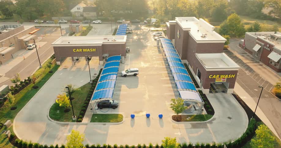 An aerial view shows a modern car wash facility with multiple bays and blue canopies under a bright morning sun