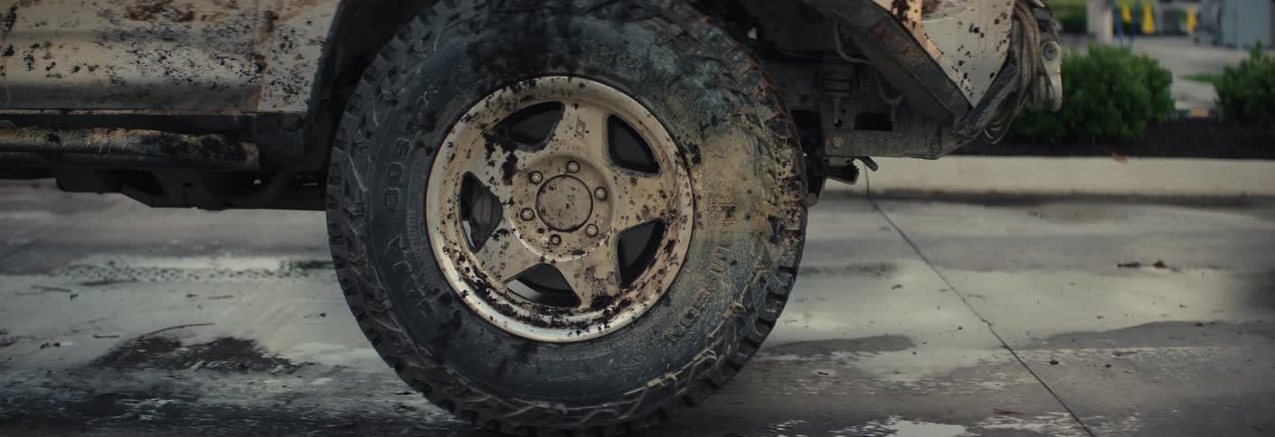 A muddy damaged pickup truck drives through a car wash exit while a building and signs sit in the background