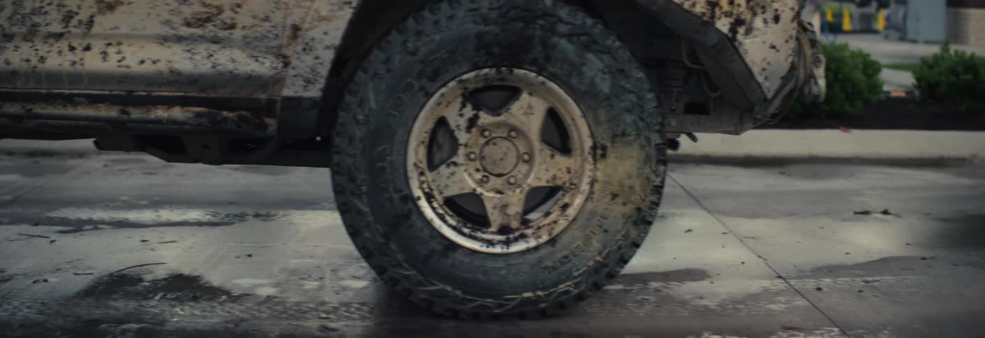 A dirty white suv covered in thick mud and debris slowly drives out of an automatic car wash bay past a brick wall and an exit sign as the camera zooms in on a mud caked tire and five spoke rim