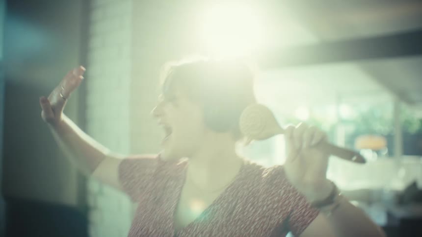 Woman wearing headphones holds a hairbrush like a microphone while singing in an indoor lit environment