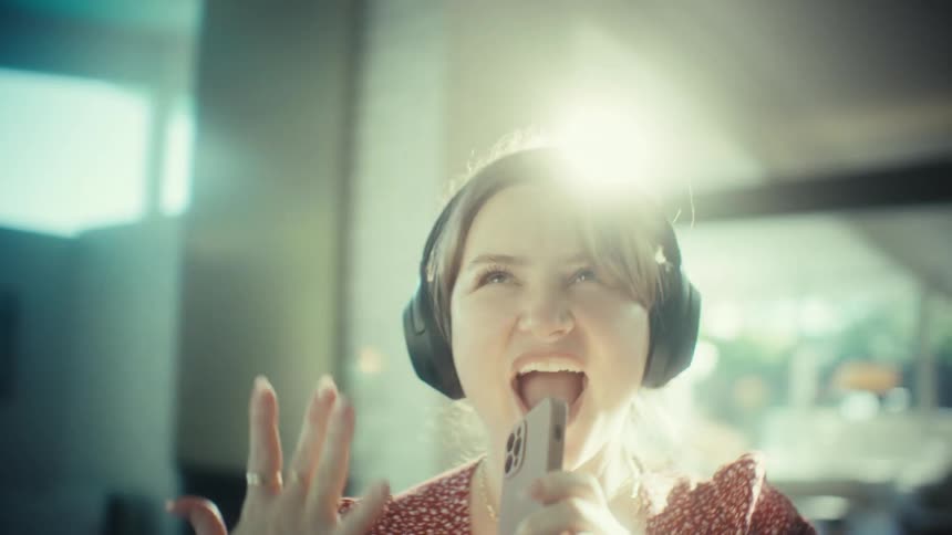 A woman wearing large black headphones and a red patterned dress dances enthusiastically in a bright roomshe sings into her smartphone as if it were a microphone smiling and gesturing with her hands while bathed in a soft warm light