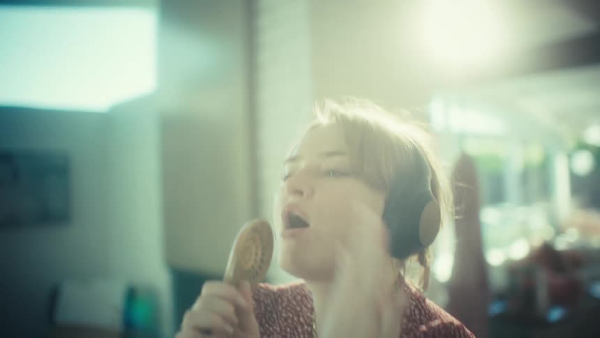 A woman wearing large black headphones and a red patterned dress sings enthusiastically into a wooden spoon while dancing in a sunlit living room with a white brick wall and modern furniture in the background